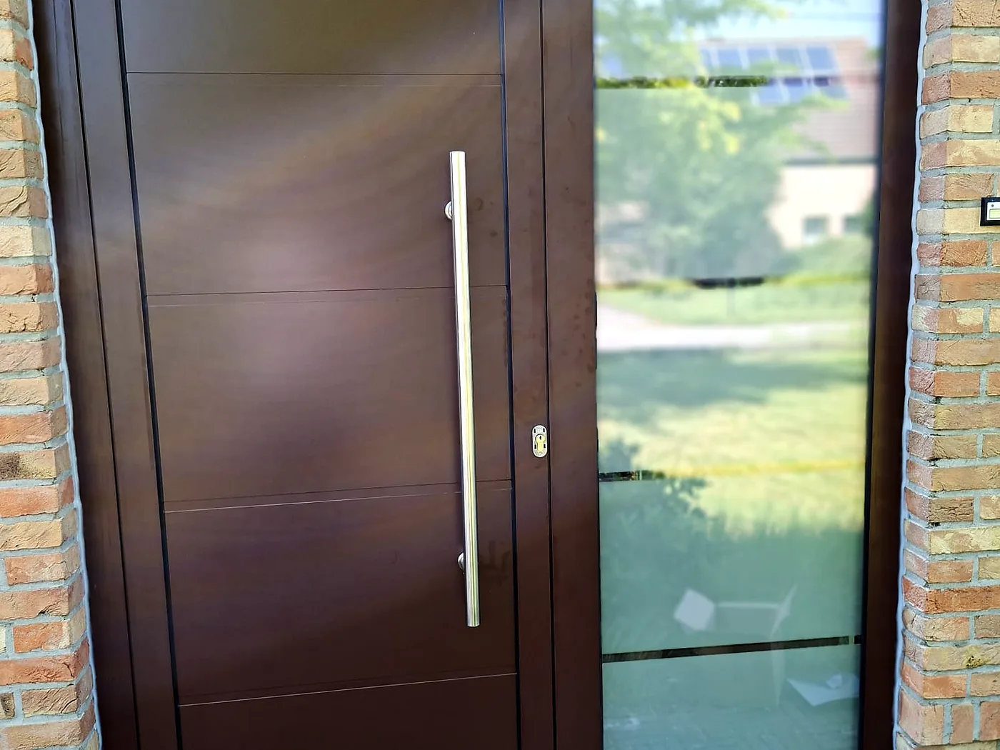 Porte d’entrée brune aux lignes modernes, longue poignée inox et vitrage latéral ouvrant sur le jardin — réalisation Pulsar Windows