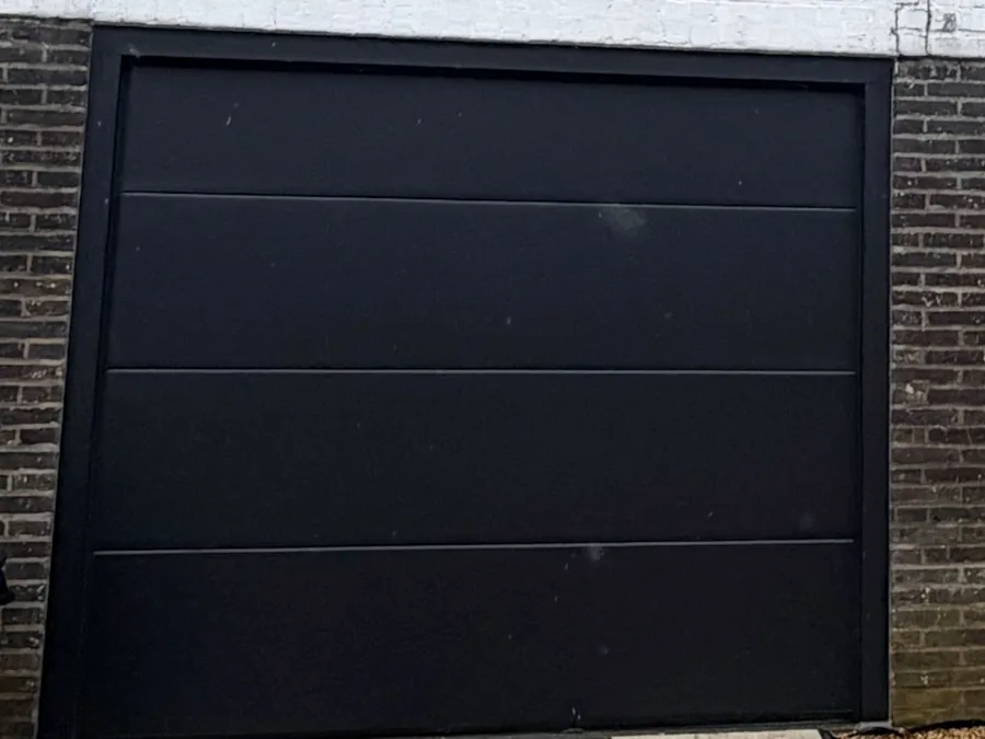 Porte de garage sectionnelle noire installée sur une façade blanche avec soubassement en briques.